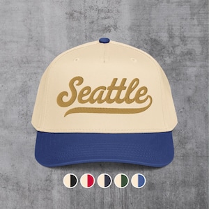 May include: A cream-coloured baseball cap with a blue brim and a small blue button on top. The front of the cap features the word Seattle embroidered in a gold-coloured script font. Below the cap are five small circular colour swatches for reference.