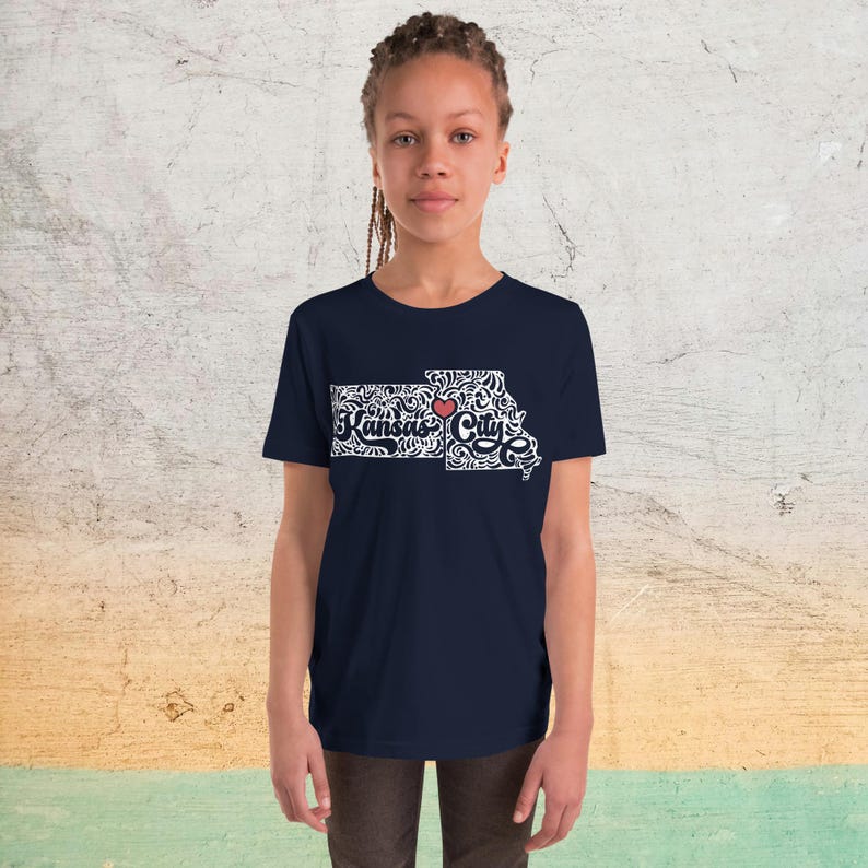 May include: A navy blue t-shirt featuring a white and red graphic of the Kansas City, Missouri, outline. The design includes the words "Kansas City" and a red heart, with decorative swirls filling the state's shape.