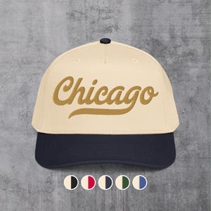 Puede incluir: Gorra de béisbol con corona color crema y visera azul marino. La palabra "Chicago" está bordada en un guion dorado en la parte delantera. La gorra tiene una visera curva y un pequeño botón azul oscuro en la parte superior.