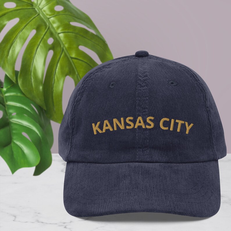 Puede incluir: Gorra de b&eacute;isbol de pana azul marino con la inscripci&oacute;n "KANSAS CITY" bordada en dorado. La gorra tiene una visera curva y un bot&oacute;n en la parte superior. El fondo incluye hojas verdes y una pared de color lila claro.