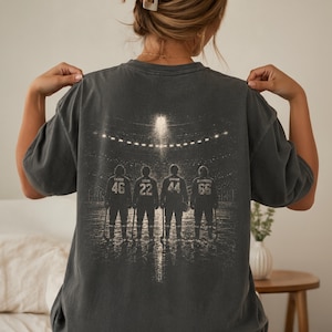 May include: A charcoal gray t-shirt featuring a back graphic of four hockey players standing on ice in a stadium. The players are numbered 46, 22, 44, and 66, with names Tucker, Logan, Graham, and Di Laurenzo printed above their respective numbers.