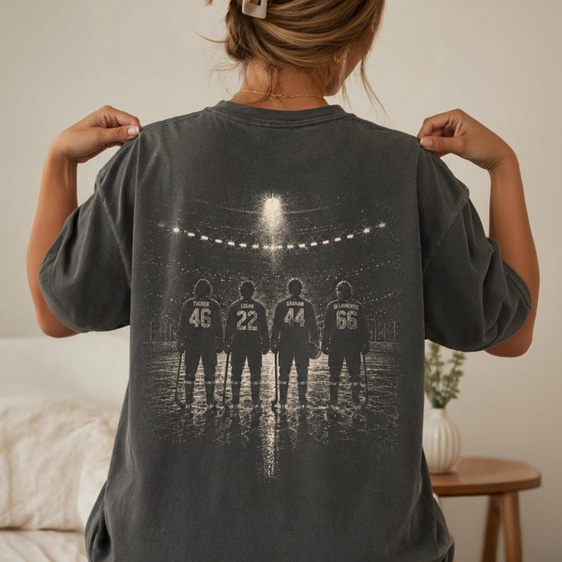 May include: A charcoal gray t-shirt featuring a back graphic of four hockey players standing on ice in a stadium. The players are numbered 46, 22, 44, and 66, with names Tucker, Logan, Graham, and Di Laurenzo printed above their respective numbers.