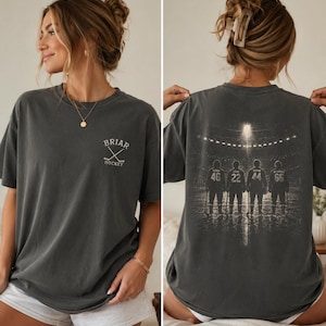 以下が含まれることがあります： チャコールグレーの半袖Tシャツ。胸元にはBRIAR HOCKEYの文字と交差したホッケースティックの小さな白いロゴがプリントされています。背面には、スタジアムの照明の下で氷上に立つ4人のホッケー選手の大きなグラフィックが描かれています。