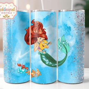 K&ouml;nnte beinhalten: Ein zylindrischer Becher mit hellblauem Aquarellhintergrund und silbernen Glitzerakzenten. Das Design zeigt eine rothaarige Meerjungfrau, die einen gelben Fisch h&auml;lt, sowie einen kleinen roten Krebs und zwei orangefarbene Fische unten.