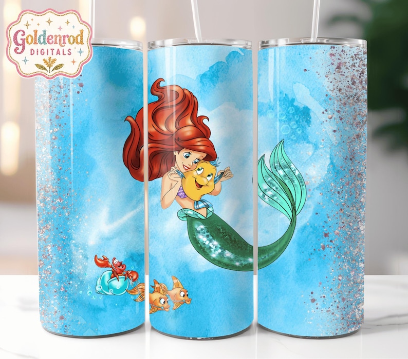 K&ouml;nnte beinhalten: Ein zylindrischer Becher mit hellblauem Aquarellhintergrund und silbernen Glitzerakzenten. Das Design zeigt eine rothaarige Meerjungfrau, die einen gelben Fisch h&auml;lt, sowie einen kleinen roten Krebs und zwei orangefarbene Fische unten.