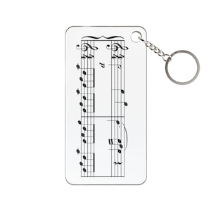 May include: A white rectangular keyring with black musical notation printed on it. The keyring has a silver-coloured chain and a round key ring attached to the top right corner. The musical notes are arranged on a staff.