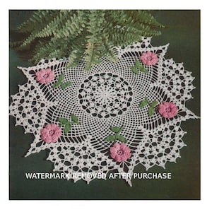 May include: White crochet doily with a delicate floral design featuring five pink flowers with green leaves. The doily is laid on a green background.