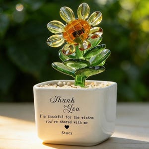 Può includere: Una scultura di girasole in vetro in un vaso di ceramica bianco. Il girasole ha petali gialli e un centro arancione. Il vaso riporta le parole "Thank You" e un messaggio, con il nome "Stacy" sotto.