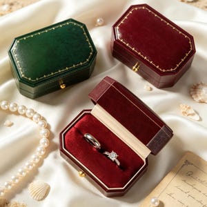 May include: Two closed octagonal jewelry boxes in dark green and deep red with gold trim, and one open deep red jewelry box containing a silver band and a silver diamond ring, accompanied by a string of white pearls and small seashells.