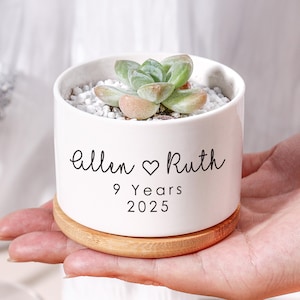 May include: A white ceramic planter with a succulent plant, resting on a wooden base. The planter has the names "Allen & Ruth", a heart symbol, and the text "9 Years 2025" in black. The succulent has green and pink leaves.