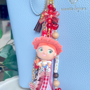 May include: A bag charm featuring a small figurine of a cowgirl with a red hat, attached to a gold-tone heart-shaped clasp. The charm includes red beads, a brown tassel, ribbons in red gingham and white lace, and beads spelling YEEHAW.