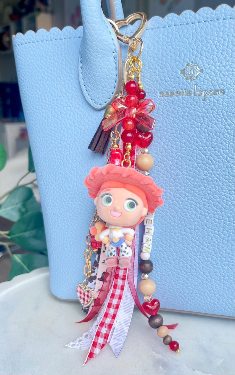 May include: A bag charm featuring a small figurine of a cowgirl with a red hat, attached to a gold-tone heart-shaped clasp. The charm includes red beads, a brown tassel, ribbons in red gingham and white lace, and beads spelling YEEHAW.