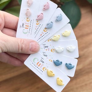 May include: A hand holds several cards displaying pairs of teapot-shaped stud earrings in various pastel colors: pink, blue, yellow, and white. The cards have the text "Too Cute clayworks" printed on them.