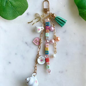 May include: A gold-colored keychain with a green tassel and various charms. Charms include a small white teapot, teacup, and plate, all with pink floral designs. The keychain spells "Spill the Tea" with colorful letter beads and is adorned with pink and pearl beads.