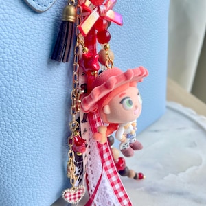 May include: A decorative bag charm featuring a miniature figurine of a character with a red hat, attached to a gold-tone chain with red beads, a small dark brown tassel, a heart-shaped charm, and various ribbons including a red and white gingham pattern.