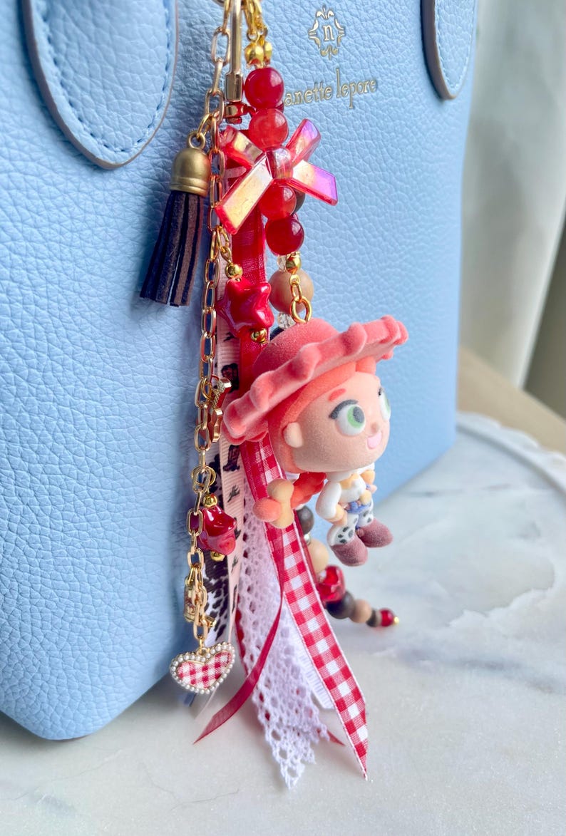 May include: A decorative bag charm featuring a miniature figurine of a character with a red hat, attached to a gold-tone chain with red beads, a small dark brown tassel, a heart-shaped charm, and various ribbons including a red and white gingham pattern.