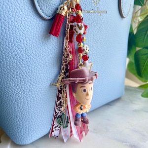 May include: A decorative bag charm featuring a miniature flocked cowboy figure, a red tassel, and various ribbons in red, white, and brown animal print. The charm includes a gold-tone chain with red, white, and gold beads, plus a small green cactus charm.