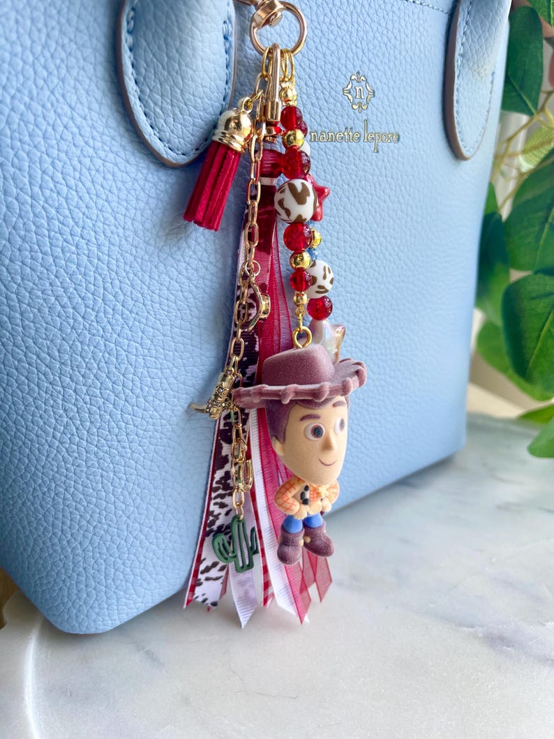 May include: A decorative bag charm featuring a miniature flocked cowboy figure, a red tassel, and various ribbons in red, white, and brown animal print. The charm includes a gold-tone chain with red, white, and gold beads, plus a small green cactus charm.