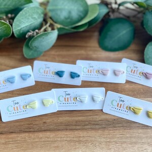 May include: A collection of colorful teacup-shaped earrings on display. Each pair is mounted on a white card with the text "Too Cute Clayworks." The earrings come in various pastel colors, including blue, pink, yellow, and white, and are arranged on a wooden surface.