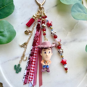 May include: A decorative keychain featuring a central figurine of a cowboy character wearing a pink hat. The accessory includes a gold heart-shaped clasp, various red and white ribbons, and dangling charms shaped like a cactus, boot, and horseshoe.