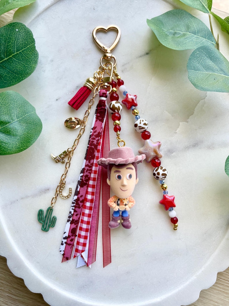 May include: A decorative keychain featuring a central figurine of a cowboy character wearing a pink hat. The accessory includes a gold heart-shaped clasp, various red and white ribbons, and dangling charms shaped like a cactus, boot, and horseshoe.