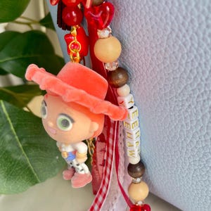 May include: A decorative bag charm featuring a small flocked character figurine wearing a coral hat, accented with red heart beads, wooden beads, and white letter beads spelling YEEHAW. The charm includes red and white gingham and lace ribbons.
