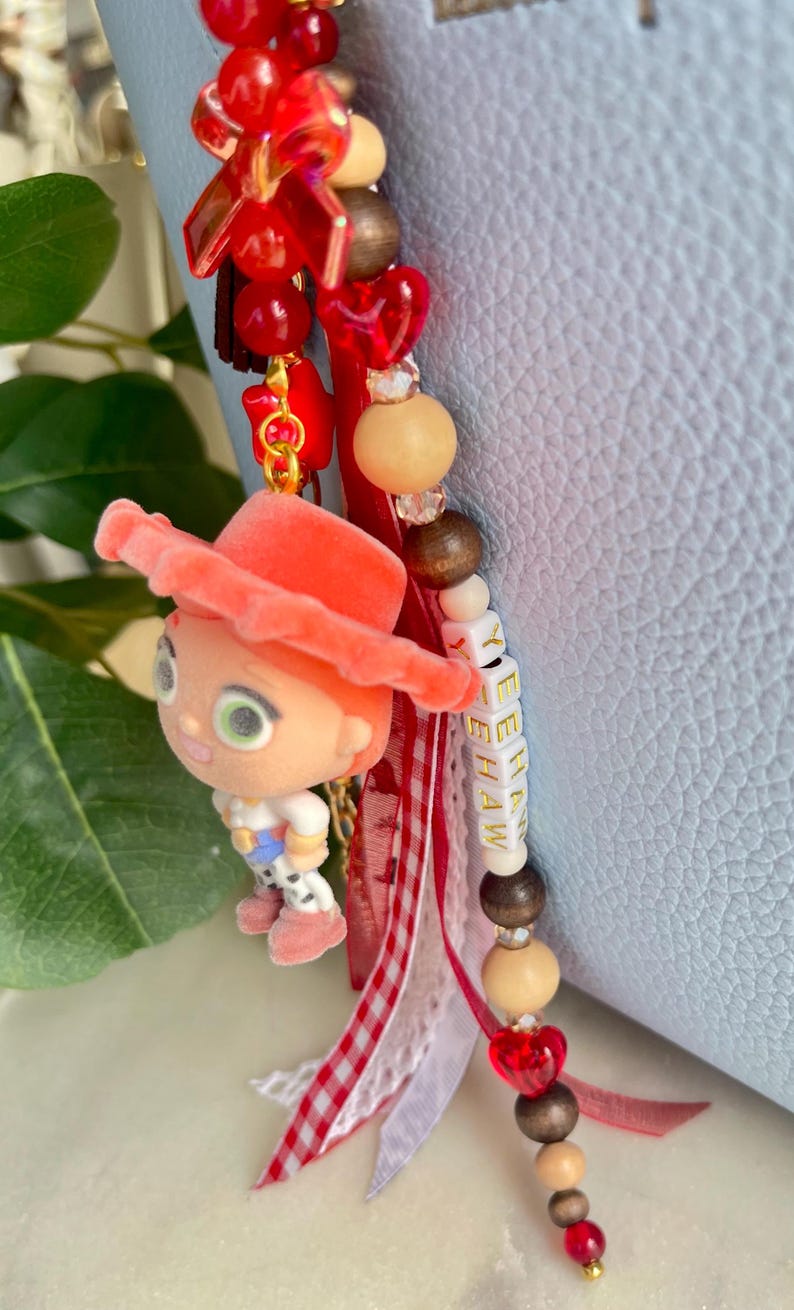 May include: A decorative bag charm featuring a small flocked character figurine wearing a coral hat, accented with red heart beads, wooden beads, and white letter beads spelling YEEHAW. The charm includes red and white gingham and lace ribbons.