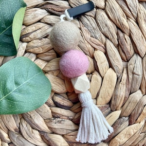 May include: An air vent car charm featuring a beige felt ball, a pink felt ball, a wooden geometric bead, and a white tassel. The charm has a black clip for attachment. The background is a woven, natural-toned material.