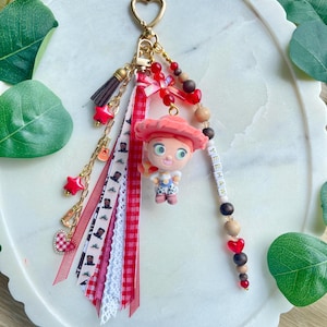 May include: A keychain featuring a gold heart-shaped clasp, a figurine of a cowgirl with red hair and a hat, and a collection of ribbons in red, white, and plaid. The accessory includes wooden and red beads, a small tassel, and the word YEEHAW.