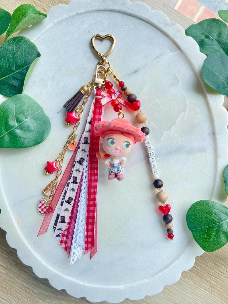 May include: A keychain featuring a gold heart-shaped clasp, a figurine of a cowgirl with red hair and a hat, and a collection of ribbons in red, white, and plaid. The accessory includes wooden and red beads, a small tassel, and the word YEEHAW.