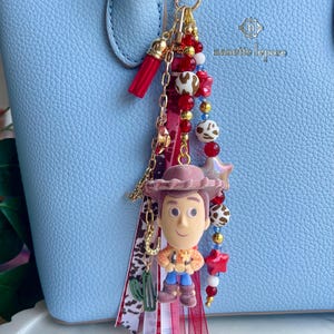 May include: A bag charm featuring a miniature Woody character figure, a red tassel, and a collection of beads in red, white, and brown cow print patterns. The charm includes gold-toned metal chains, a small green cactus charm, and a horseshoe charm, all attached to a heart-shaped gold clasp.