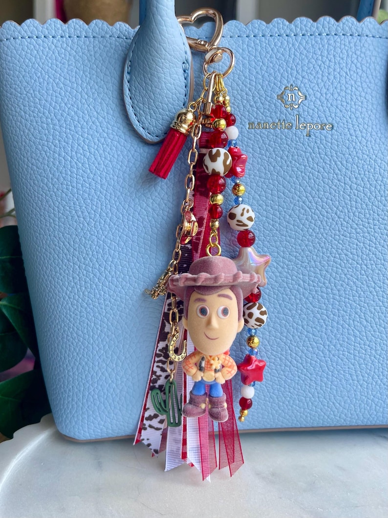 May include: A bag charm featuring a miniature Woody character figure, a red tassel, and a collection of beads in red, white, and brown cow print patterns. The charm includes gold-toned metal chains, a small green cactus charm, and a horseshoe charm, all attached to a heart-shaped gold clasp.