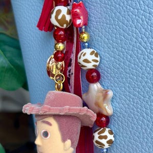 May include: A bag charm featuring a miniature flocked cowboy figure, red and white cow-print beads, red star-shaped beads, gold-tone metallic beads, and a red fabric tassel. The charm is attached to a light blue textured handbag with the text nanette lepore.