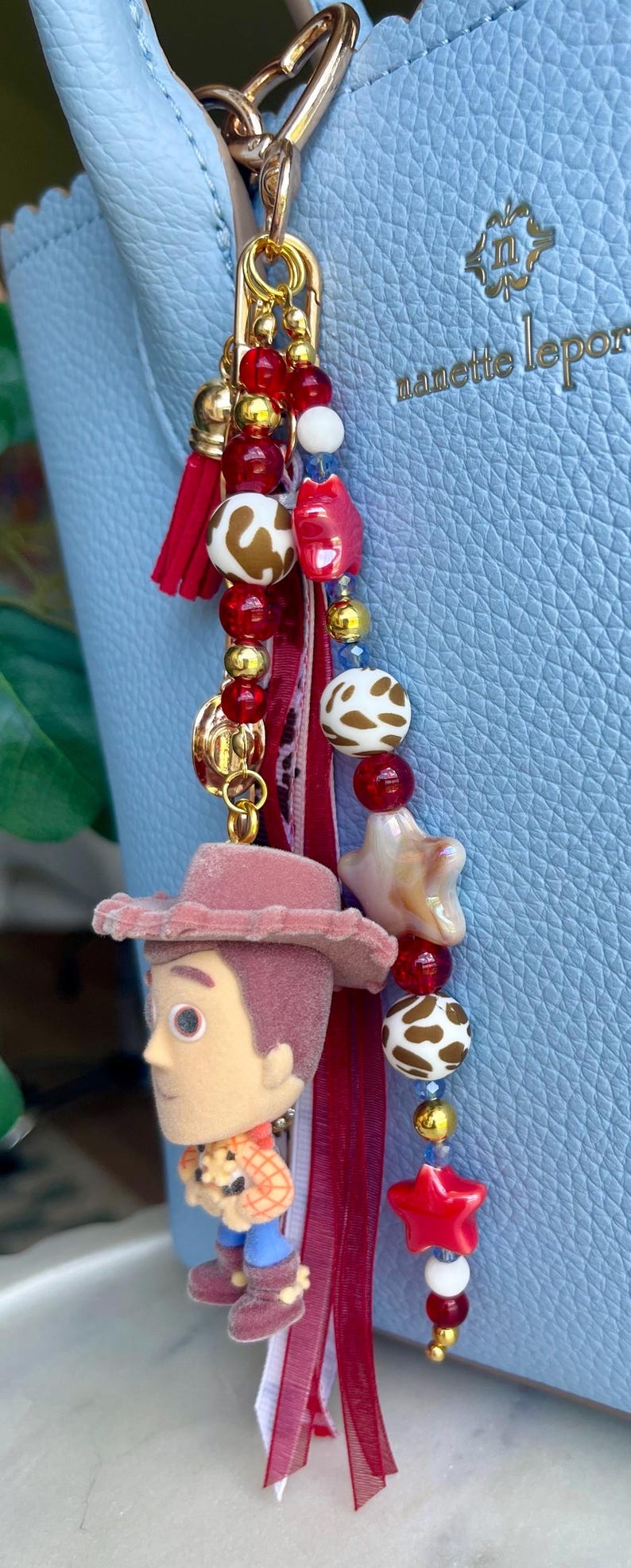 May include: A bag charm featuring a miniature flocked cowboy figure, red and white cow-print beads, red star-shaped beads, gold-tone metallic beads, and a red fabric tassel. The charm is attached to a light blue textured handbag with the text nanette lepore.