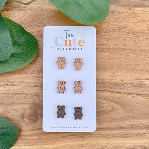 May include: A white card displays four pairs of bear-shaped earrings in various shades of brown. The card reads "Too Cute clayworks" in colorful text. The earrings are arranged in two rows of two, with different patterns and colors.