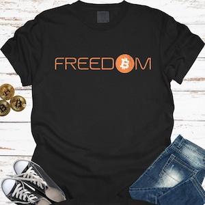 May include: Black t-shirt with the word "FREEDOM" in orange, featuring a Bitcoin symbol. The shirt is paired with blue jeans and black and white sneakers. Bitcoin coins are visible on the left.