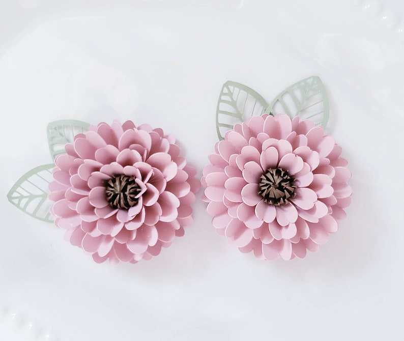 May include: Two pink paper flowers with green paper leaves. The flowers have a layered, ruffled appearance and a brown center.