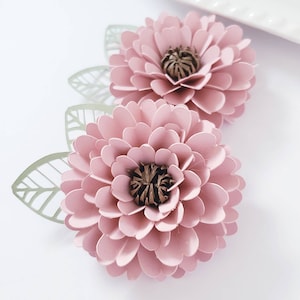 May include: Two pink paper flowers with brown centers and green paper leaves. The flowers are layered and have a delicate, feminine look.