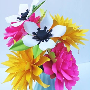 May include: A bouquet of paper flowers in a light blue vase. The arrangement includes white anemones with black centers, bright pink peonies, and yellow dahlias. Green leaves and accents add to the colorful display.