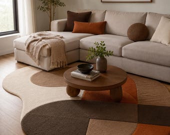 May include: A large, irregularly shaped area rug featuring a geometric pattern in shades of cream, tan, brown, and burnt orange. The rug has a textured, tufted surface and is positioned under a round wooden coffee table with thick, rounded legs.