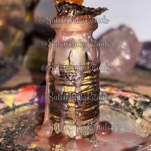 Może przedstawiać: Dekoracyjna szklana butelka ze złotym i brązowym wzorem, pokryta różowym woskiem. Butelka ma ciemną górę i jest otoczona różowym płynem. Tekst "@SalemBabyRituals" jest powtórzony na butelce.