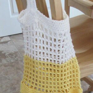 May include: A handmade, open-weave crochet tote bag in white and yellow. The bag is hanging on a wooden chair. The top portion is white, transitioning to a yellow base. The bag's open weave design is visible.