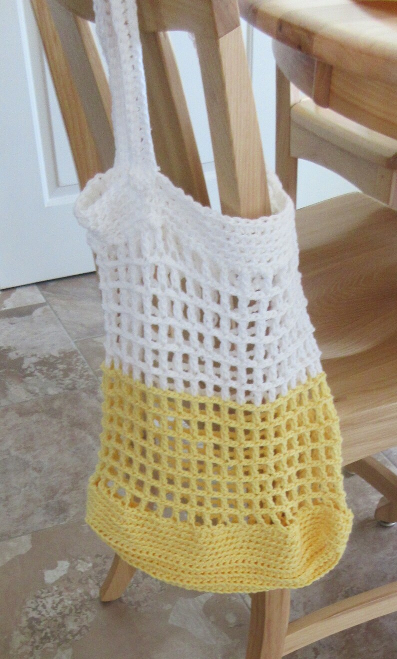 May include: A handmade, open-weave crochet tote bag in white and yellow. The bag is hanging on a wooden chair. The top portion is white, transitioning to a yellow base. The bag's open weave design is visible.