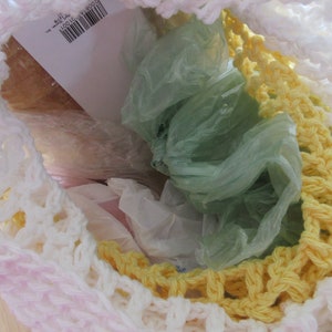 May include: A handmade crochet bag in white, yellow, and pink, filled with plastic bags. The bag's open weave reveals the contents, with a barcode label visible inside. The crochet work features a textured pattern.