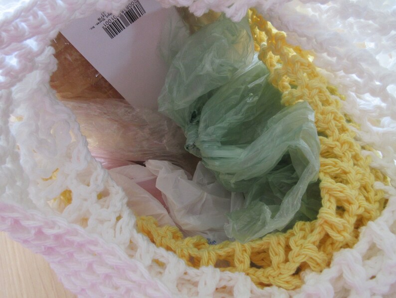 May include: A handmade crochet bag in white, yellow, and pink, filled with plastic bags. The bag's open weave reveals the contents, with a barcode label visible inside. The crochet work features a textured pattern.