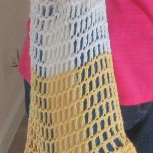 May include: A handmade crochet tote bag in white, yellow, and a hint of blue. The bag is open-weave, with a rounded bottom and a long strap. The bag is held by a person wearing a pink shirt and blue jeans.