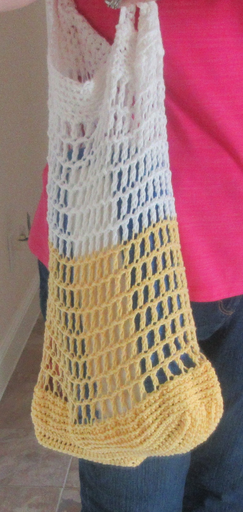 May include: A handmade crochet tote bag in white, yellow, and a hint of blue. The bag is open-weave, with a rounded bottom and a long strap. The bag is held by a person wearing a pink shirt and blue jeans.