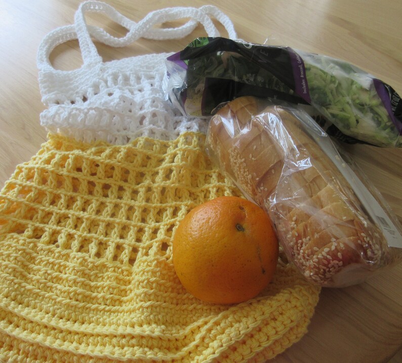 May include: A handmade crochet shopping bag in white and yellow, filled with groceries. The bag has a white top and yellow body. An orange, a loaf of bread, and a bag of lettuce are visible.