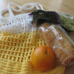 May include: A handmade crochet tote bag in white and yellow, filled with groceries. The bag holds a loaf of bread, a bag of salad, and an orange. The bag is made of a sturdy, open weave design.