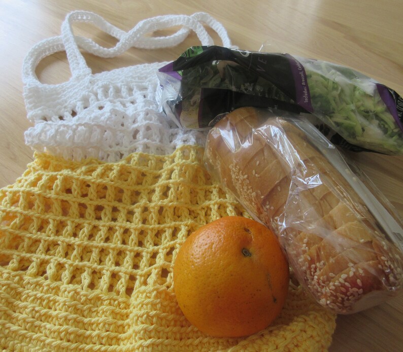 May include: A handmade crochet tote bag in white and yellow, filled with groceries. The bag holds a loaf of bread, a bag of salad, and an orange. The bag is made of a sturdy, open weave design.
